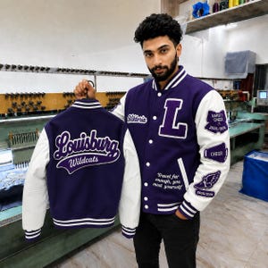 Könnte beinhalten: Eine violett-weiße Collegejacke mit dem Schriftzug Louisburg Wildcats auf der Rückseite, dem Buchstaben L auf der Vorderseite und dem Zitat Travel far enough, you meet yourself. Die Ärmel sind weiß und mit Varsity Cheer Aufnähern versehen.