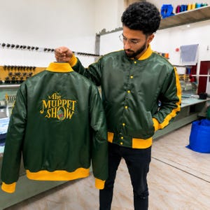 Könnte beinhalten: Eine dunkelgrüne Satin-Bomberjacke mit gelben Akzenten an Kragen, Ärmelbündchen und Saum. Auf der Rückseite befindet sich ein gelb gesticktes Logo mit der Aufschrift The Muppet Show und einer kleinen grünen Grafik. Mit Druckknopfverschluss.