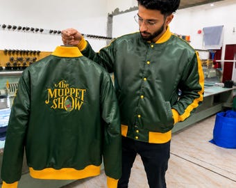 May include: A dark green satin bomber jacket with yellow trim on the collar, cuffs, and hem. The back features an embroidered yellow logo that reads The Muppet Show with a small green character graphic. The jacket has a snap-button front closure.