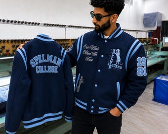 May include: A navy blue varsity jacket with light blue striped trim on the collar, cuffs, and hem. The back features the text "SPELMAN COLLEGE" in light blue. The front includes "Our Whole School For Trust", "SPELMAN JAGUARS", and "1981".