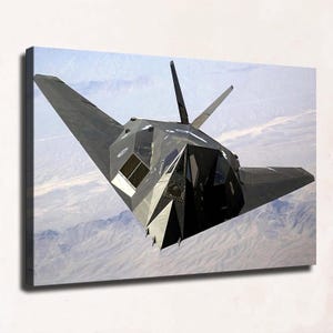 May include: A black F-117 Nighthawk stealth aircraft flying over a mountainous landscape. The jet features a distinct angular, faceted design with sharp geometric lines, a pointed nose, and V-shaped tail fins, captured in mid-flight.
