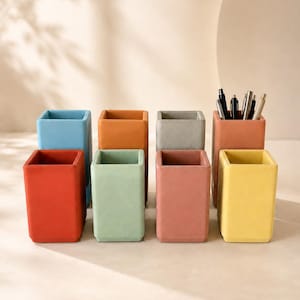 May include: A collection of eight rectangular pencil holders in various solid matte colours including red, light blue, orange, light green, grey, terracotta, and yellow, with one terracotta holder containing several pens and pencils.