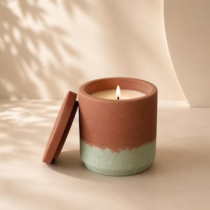 May include: A lit candle in a cylindrical container made of two-toned material, featuring a terracotta-colored upper section and a pale sage green lower section. A matching terracotta-colored circular lid rests upright against the side of the candle.