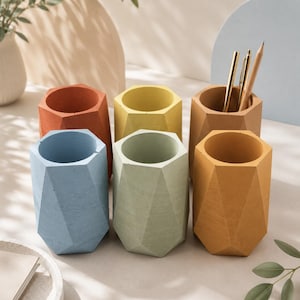 Op de afbeelding: Een verzameling van zes zeshoekige, geometrisch gevormde bureau-organizers van mat materiaal in diverse kleuren, waaronder terracotta, geel, lichtblauw, saliegroen, mosterdgeel en beige, met metalen pennen in een van de houders.