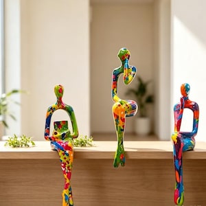 May include: Three abstract human-shaped sculptures in a sitting pose, each featuring a vibrant, multi-colored paint splatter pattern with hues of blue, red, yellow, green, and orange. The figures are positioned on a flat, light-toned wooden surface.