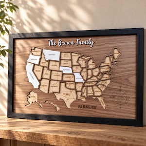 May include: A framed wooden wall map of the USA featuring individual state cutouts, with some states highlighted in white. The text "The Brown Family" appears at the top, and "USA TRAVEL MAP" is printed at the bottom right with a compass rose.
