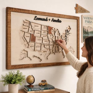 Peut inclure: Une carte murale en bois des États-Unis portant les noms Leonard + Amelia en haut. La carte se compose de pièces d'État individuelles, certaines étant mises en évidence par un ton de bois plus foncé. Une rose des vents est visible en bas à droite.