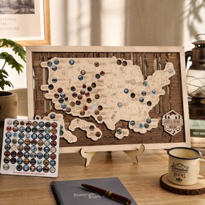 Puede incluir: Un mapa de madera de los Estados Unidos con recortes para parques nacionales, en un marco de madera clara. El mapa incluye fichas circulares, una hoja separada de pegatinas de parques y una placa hexagonal con el texto James & Sophia Travel Map.