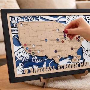 May include: A framed wooden map of the United States featuring cutouts for baseball stadiums. The map includes text labels for various ballparks and the words BASEBALL STADIUM MAP. The design incorporates blue and white layered accents.