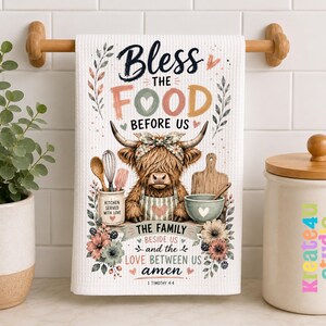 Puede incluir: Un paño de cocina blanco con textura de gofre que presenta una ilustración central de una vaca de las Tierras Altas con una diadema floral. El texto dice Bless the food before us, the family beside us and the love between us amen, 1 Timothy 4:4.