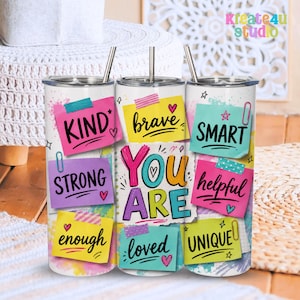 Op de afbeelding: Drie cilindrische bekers met metalen rietjes, met een witte achtergrond en kleurrijke plakbriefjes. De briefjes tonen de woorden KIND, brave, SMART, STRONG, YOU ARE, helpful, enough, loved en UNIQUE in een zwart handschrift.