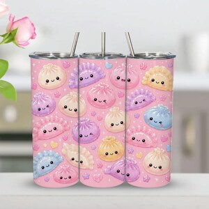 May include: A cylindrical tumbler with a light pink background featuring a repeating pattern of various colorful, cartoon-style dumplings with smiling faces, small hearts, and flowers. The tumbler includes a clear lid and a thin metal straw.
