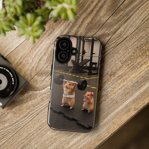 May include: A smartphone case featuring a graphic of two hamsters in a gym. One hamster wears a white weightlifting belt and lifts a barbell with black plates. The other hamster stands nearby wearing pink wristbands and a small pink bow on its head.