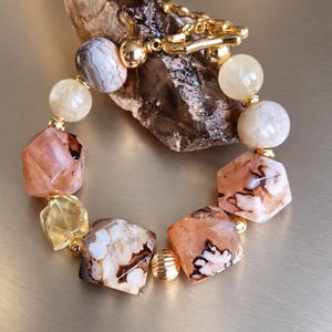 May include: A beaded bracelet featuring faceted peach-colored stones with dark brown inclusions, smooth translucent yellow beads, a grey-banded agate bead, and small gold-toned metal spacers. The bracelet includes a gold-toned toggle clasp closure.