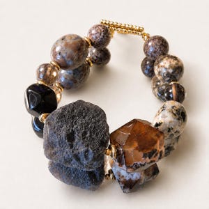 May include: A bracelet featuring a mix of smooth, polished, and rough-textured beads in shades of brown, black, and cream. The beads are accented with small gold-toned metal spacers and a gold-toned toggle clasp, creating a varied, multi-textured design.