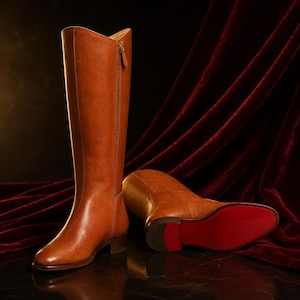 Puede incluir: Un par de botas altas de cuero color canela con acabado liso. Una bota está de pie, con una cremallera lateral y una pequeña lengüeta, mientras que la otra está tumbada, mostrando una suela roja distintiva y un tacón bajo y apilado.