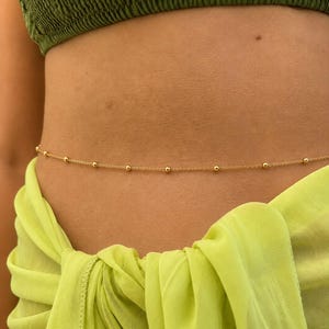 May include: A delicate gold-toned waist chain featuring evenly spaced small round beads along a thin metal link chain. The jewelry rests horizontally across the midsection, positioned between a dark olive green top and a bright lime green sarong.