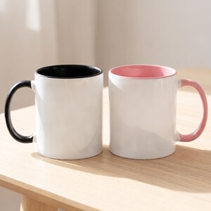 May include: Two ceramic mugs with white exterior bodies. The mug on the left features a solid black interior and a black handle. The mug on the right features a solid light pink interior and a matching light pink handle. Both mugs are cylindrical.