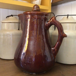 May include: A brown glazed ceramic pitcher with a wooden handle and a lid. The pitcher is sitting on a wooden surface next to two white ceramic jars with lids.