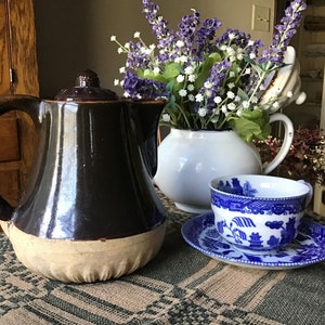 May include: A brown and white glazed teapot with a handle, a white teapot with a handle, a blue and white teacup and saucer, and a bouquet of purple and white flowers in a white vase.