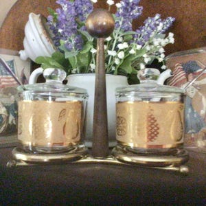 May include: Two glass jars with gold lids and a gold design on the sides, sitting on a gold metal stand. The jars are filled with a clear liquid. The stand has a round top and three legs. The jars are on a wooden surface.