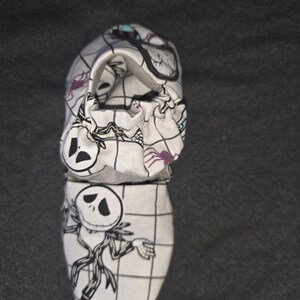 May include: A white fabric baby shoe featuring a black grid pattern and illustrations of a skeletal character with a round head and thin limbs. The shoe has an elasticized opening and includes small purple spider motifs printed on the fabric.
