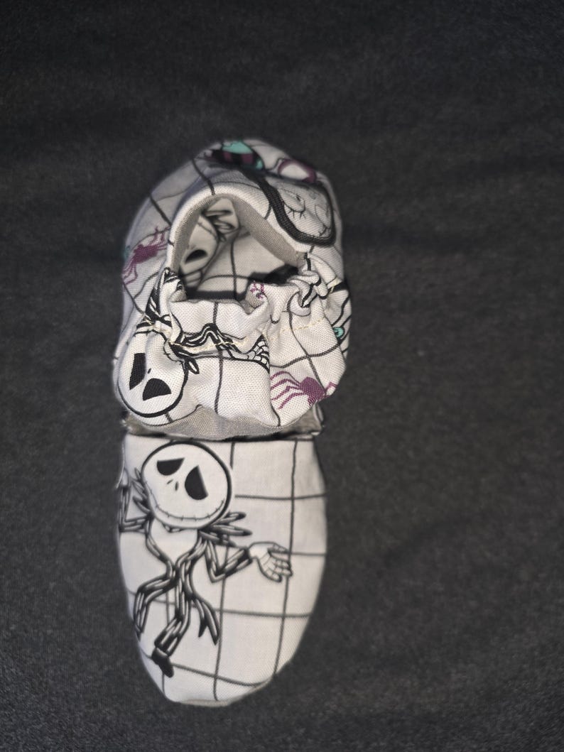 May include: A white fabric baby shoe featuring a black grid pattern and illustrations of a skeletal character with a round head and thin limbs. The shoe has an elasticized opening and includes small purple spider motifs printed on the fabric.