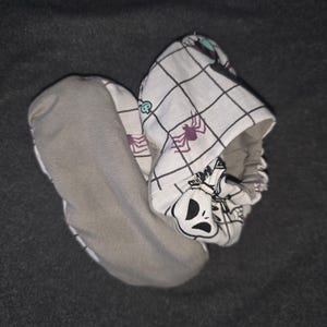 May include: A pair of soft baby booties featuring a white fabric with a black grid pattern, purple spider illustrations, and a character face graphic. The soles are made of a solid grey fabric, and the openings have an elasticized gathered edge.