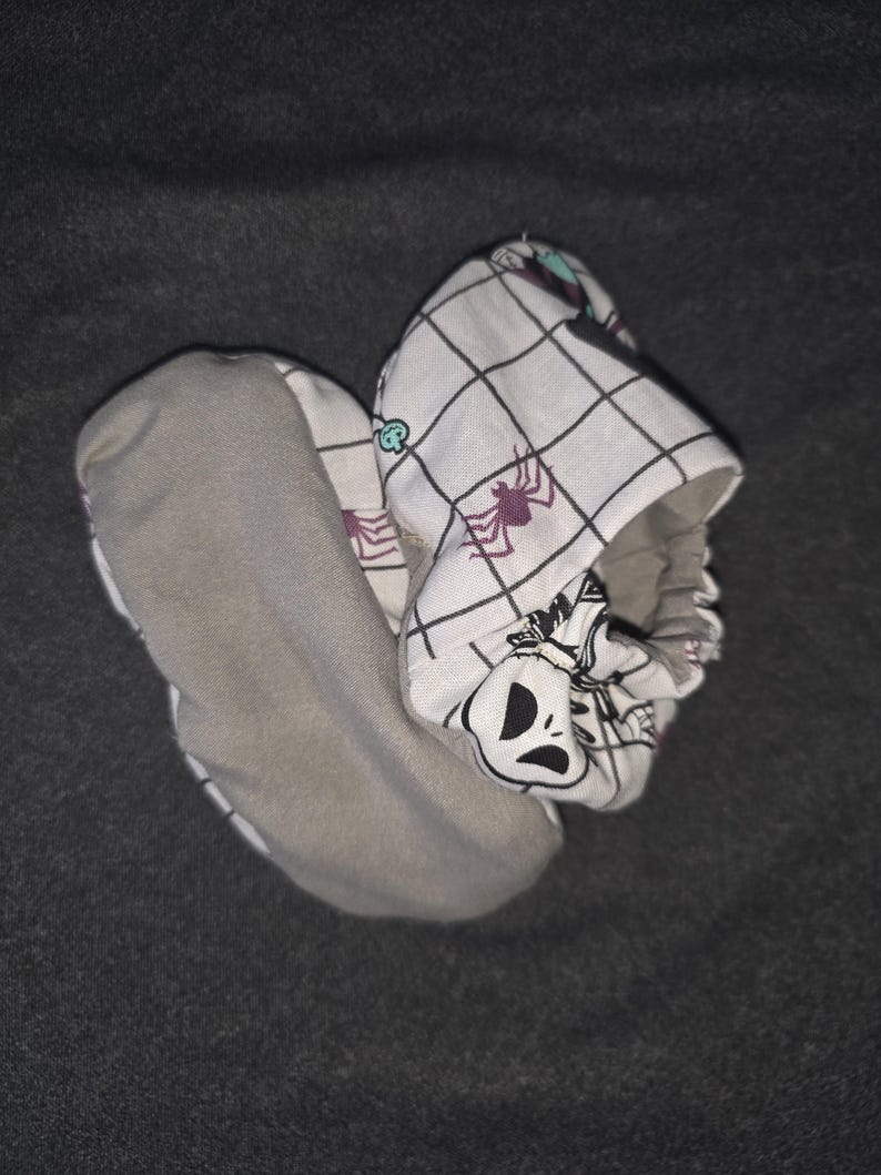May include: A pair of soft baby booties featuring a white fabric with a black grid pattern, purple spider illustrations, and a character face graphic. The soles are made of a solid grey fabric, and the openings have an elasticized gathered edge.