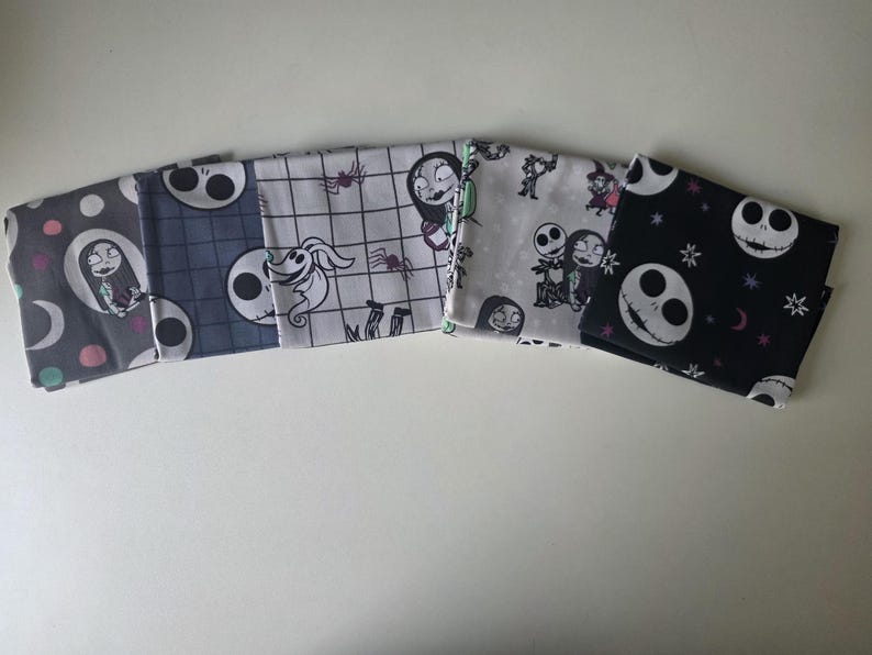 May include: Five folded fabric squares featuring characters and motifs from The Nightmare Before Christmas. Patterns include Sally on a gray background, Jack Skellington on blue plaid, Zero on a grid, various characters on white, and Jack on black.
