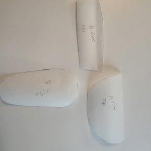 May include: Three white paper pattern pieces for footwear construction. Each piece has hand-written text in black ink. The pieces are labeled 12-15mo heel, 12-15mo side, and 12-15mo top. The shapes are cut out with visible black outlines.