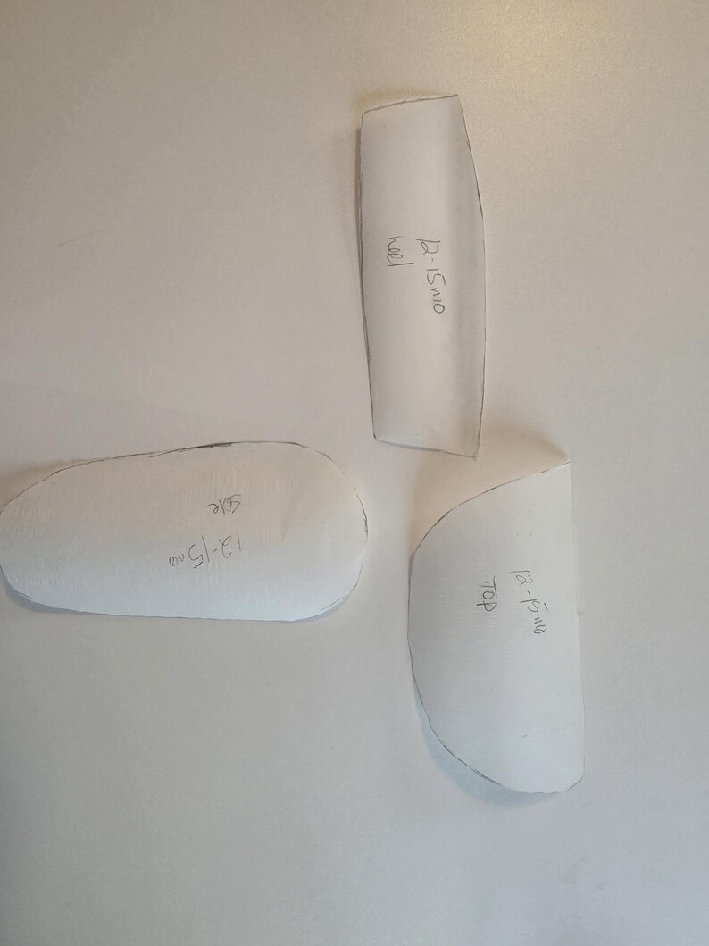 May include: Three white paper pattern pieces for footwear construction. Each piece has hand-written text in black ink. The pieces are labeled 12-15mo heel, 12-15mo side, and 12-15mo top. The shapes are cut out with visible black outlines.