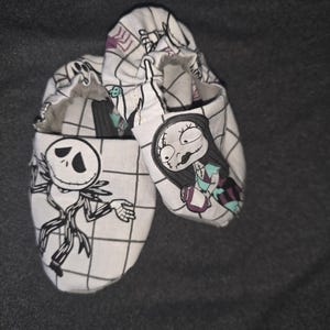May include: A pair of soft-soled baby shoes featuring a white fabric with a black grid pattern. One shoe displays a black and white illustration of Jack Skellington, while the other shoe features a colorful illustration of Sally from The Nightmare Before Christmas.