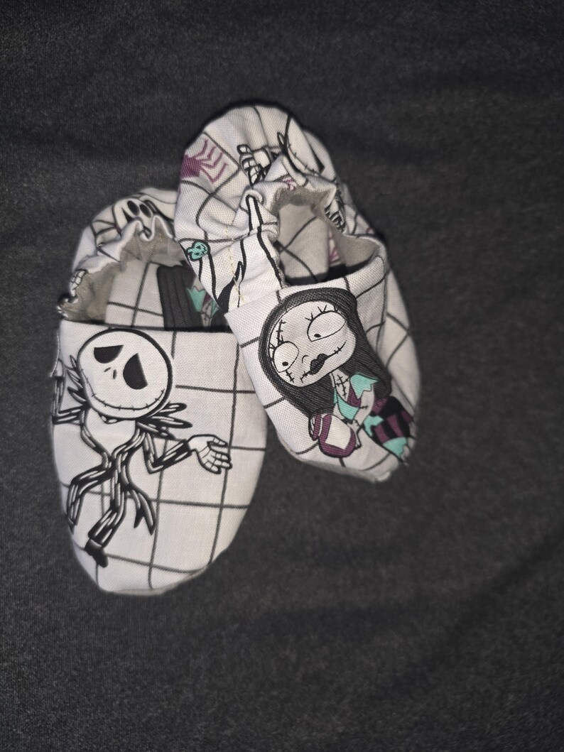 May include: A pair of soft-soled baby shoes featuring a white fabric with a black grid pattern. One shoe displays a black and white illustration of Jack Skellington, while the other shoe features a colorful illustration of Sally from The Nightmare Before Christmas.