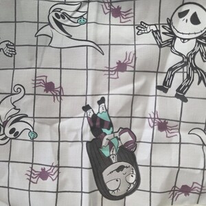 May include: A white fabric featuring a black grid pattern with repeating illustrations of Jack Skellington, Zero the ghost dog, and Sally. The design includes small purple spiders scattered throughout the grid, creating a detailed, repeating character print.