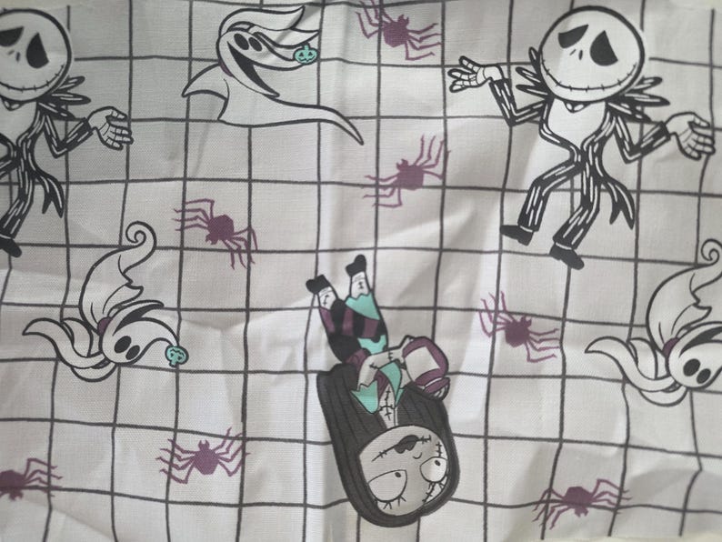 May include: A white fabric featuring a black grid pattern with repeating illustrations of Jack Skellington, Zero the ghost dog, and Sally. The design includes small purple spiders scattered throughout the grid, creating a detailed, repeating character print.