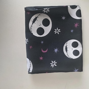 May include: A folded piece of black fabric featuring a repeating pattern of white skull faces with stitched mouths, interspersed with small white and purple stars and crescent moons. The design is printed across the dark background material.