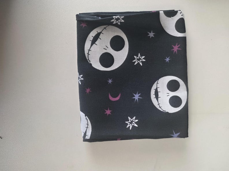 May include: A folded piece of black fabric featuring a repeating pattern of white skull faces with stitched mouths, interspersed with small white and purple stars and crescent moons. The design is printed across the dark background material.