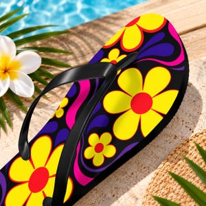 May include: A flip-flop featuring a black base with a vibrant pattern of large yellow flowers with red centers, interspersed with purple and pink wavy shapes. The footwear includes a smooth black rubber strap attached to the sole.