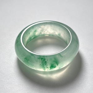 May include: A smooth, polished band ring crafted from translucent light green stone with darker green mottled inclusions throughout the material. The ring features a rounded, thick profile with a glossy finish that reflects light across its surface.