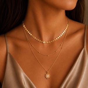 Puede incluir: Un conjunto de collar de tres capas en oro que presenta una cadena superior con pequeños discos circulares planos, una cadena intermedia con pequeñas cuentas doradas espaciadas y una cadena inferior con un colgante de sol con piedra blanca.