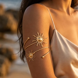 May include: A gold-toned metal arm cuff featuring a spiral design that wraps around the upper arm. The piece is adorned with two sun-shaped motifs, each with a central circular disc and radiating wavy rays, finished with small spherical end caps.
