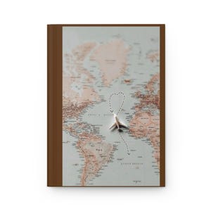 May include: A hardcover journal featuring a world map on the cover with a muted color palette. A small white airplane figurine is positioned in the center, with a dashed line forming a heart shape around it, tracing a flight path across the ocean.