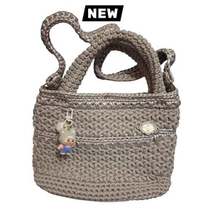 Può includere: Una borsa all'uncinetto color tortora metallizzato con corpo rettangolare strutturato, due manici e una piccola placca in metallo argentato sul davanti. Un piccolo ciondolo a forma di personaggio dei cartoni animati pende da una chiusura argentata ed è visibile un'etichetta nera con il testo NEW.