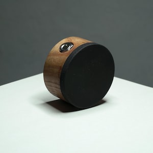 May include: A cylindrical object featuring a natural light brown wood grain body and a solid matte black circular top. A small, circular silver metal port or input jack is embedded into the side of the wooden section of the cylindrical component.