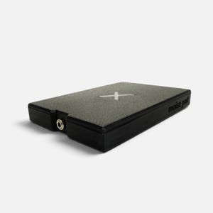 Op de afbeelding: Een rechthoekig zwart elektronisch apparaat met een grijs gestructureerd bovenoppervlak voorzien van een wit X-symbool. De zijkant van het apparaat is gelabeld met de tekst "noise pad" en bevat een ronde metalen ingangspoort aan de voorzijde.