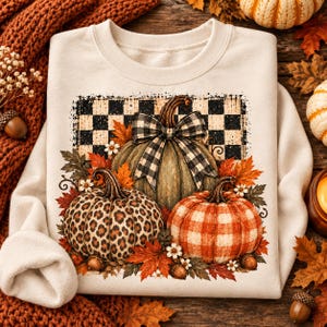 May include: A cream-colored crewneck sweatshirt featuring a graphic print of three pumpkins. The center pumpkin has a black and white buffalo check bow, the left has a leopard print, and the right has an orange plaid pattern, all set against a black and white checkered background with autumn leaves.