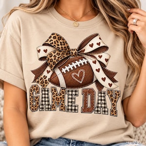 Op de afbeelding: Een beige t-shirt met een afbeelding van een bruine football achter een grote strik met luipaardprint en witte hartjes. Onder de football staat de tekst "GAME DAY" in grote blokletters met luipaard- en ruitpatronen op de letters gedrukt.