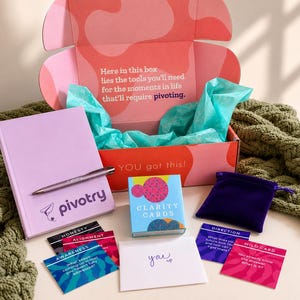 May include: A gift box with pink and orange wavy patterns containing turquoise tissue paper, a light purple journal with a matching pen, a deck of Clarity Cards, a small purple velvet pouch, and several colourful prompt cards with text.
