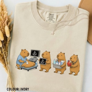 Puede incluir: Una camiseta color marfil con un gráfico de cuatro capibaras como personal médico. Uno realiza una ecografía, otro sostiene una imagen, el tercero lleva suministros médicos y el cuarto sostiene un modelo de corazón con un estetoscopio.